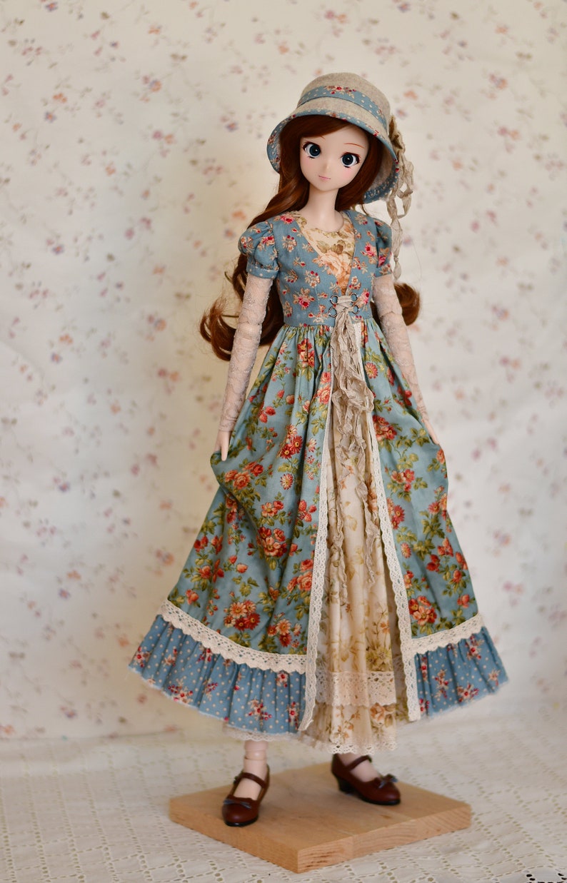 May include: A doll wearing a blue floral print dress with a matching hat. The dress features a V-neck, lace trim, and a ruffled front. The doll has long brown hair and is wearing brown shoes. The background is a floral pattern.