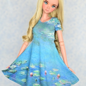 May include: A doll wearing a blue dress with a water lily print. The dress has short sleeves and a V-neck. The doll has long blonde hair and blue eyes. The dress features a Monet-style water lily design.