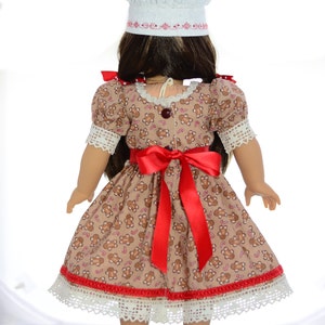 May include: A doll wearing a gingerbread-print dress with a red ribbon, white lace trim, and a white chef's hat. The doll has brown hair and is wearing white shoes.