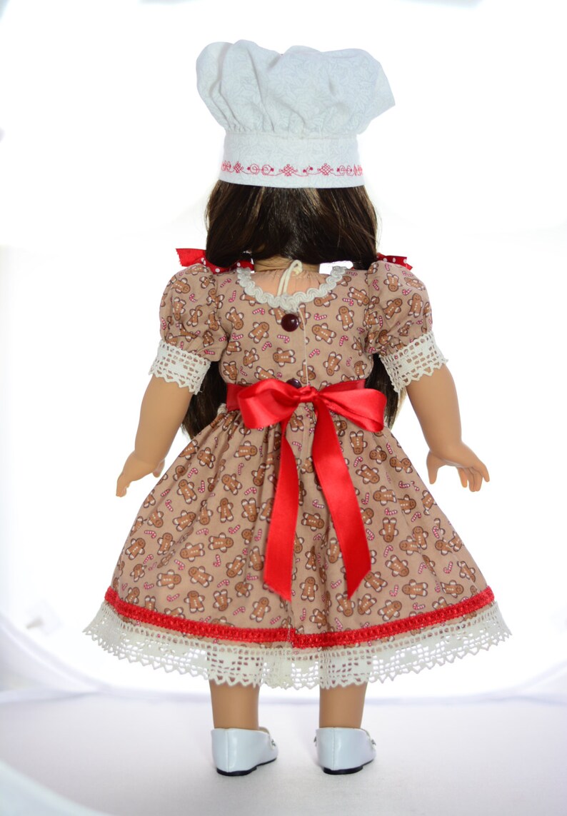 May include: A doll wearing a gingerbread-print dress with a red ribbon, white lace trim, and a white chef's hat. The doll has brown hair and is wearing white shoes.