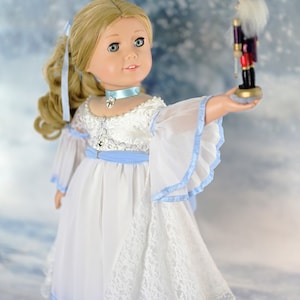 May include: A doll wearing a white dress with blue trim and a lace overlay, holding a wooden nutcracker toy. The doll has blonde hair and blue eyes. The background is a snowy scene.
