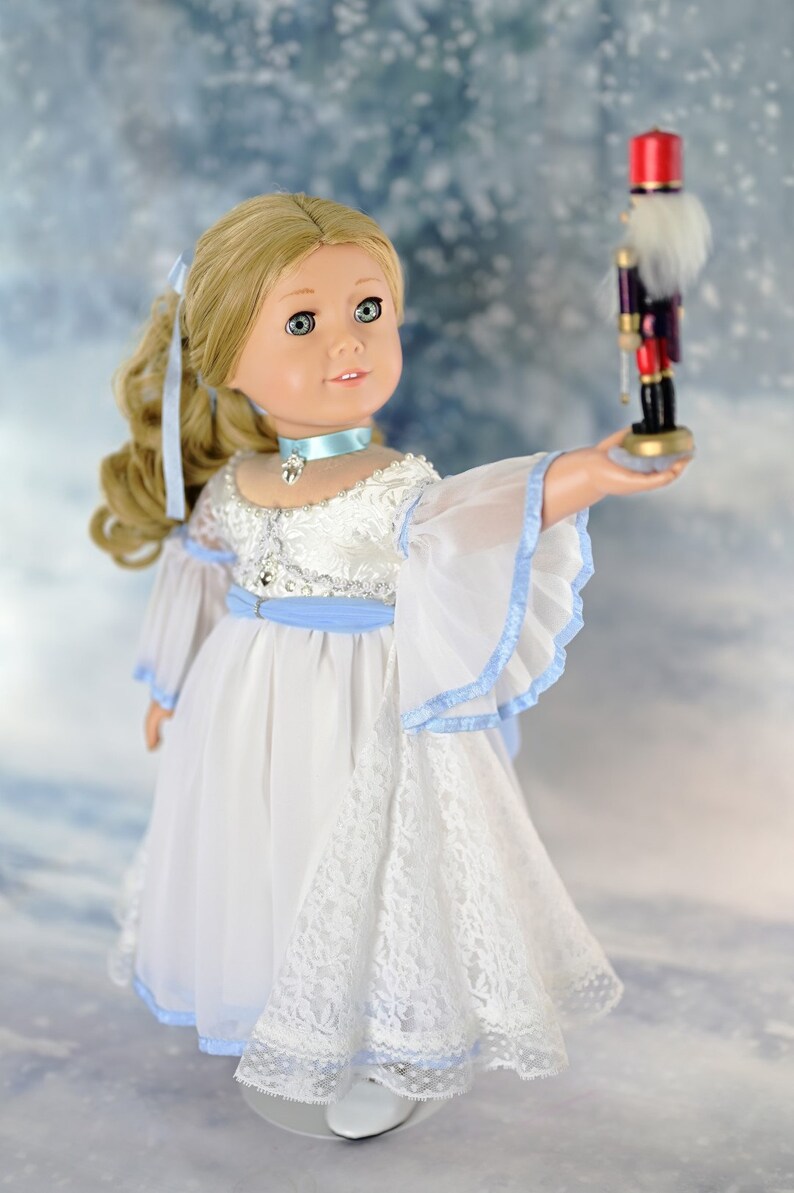 May include: A doll wearing a white dress with blue trim and a lace overlay, holding a wooden nutcracker toy. The doll has blonde hair and blue eyes. The background is a snowy scene.