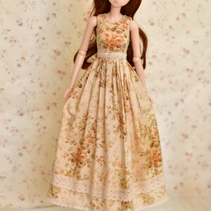 May include: A doll wearing a sleeveless, floral-print dress with a lace trim at the waist and hem. The dress features a beige background with orange and brown roses. The doll has long brown hair and is wearing brown shoes.