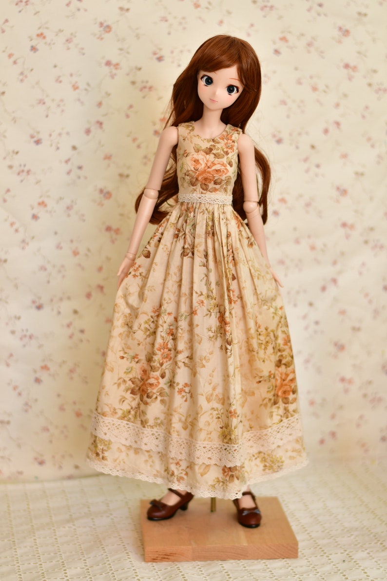May include: A doll wearing a sleeveless, floral-print dress with a lace trim at the waist and hem. The dress features a beige background with orange and brown roses. The doll has long brown hair and is wearing brown shoes.