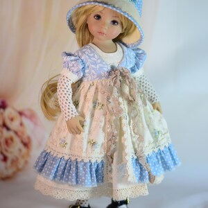 K&ouml;nnte beinhalten: Porzellanpuppe mit langem blondem Haar, gekleidet in einem blau-wei&szlig; gebl&uuml;mten Kleid, passendem Hut und schwarzen Schuhen. Das Kleid hat Spitzen- und R&uuml;schendetails. Die Puppe ist ein Sammlerst&uuml;ck.