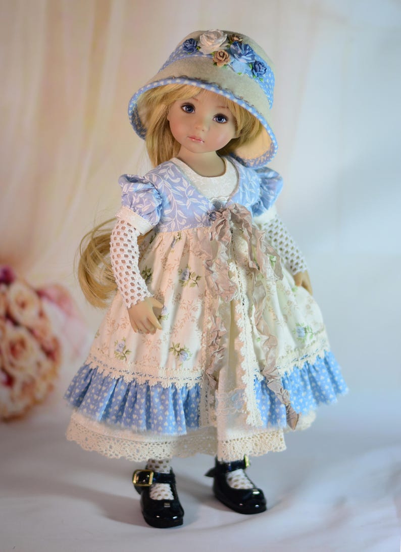 K&ouml;nnte beinhalten: Porzellanpuppe mit langem blondem Haar, gekleidet in einem blau-wei&szlig; gebl&uuml;mten Kleid, passendem Hut und schwarzen Schuhen. Das Kleid hat Spitzen- und R&uuml;schendetails. Die Puppe ist ein Sammlerst&uuml;ck.