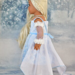 May include: A doll wearing a light blue dress with a white lace overlay and a white fur trim on the sleeve. The doll has long blonde hair and is wearing a silver crown.