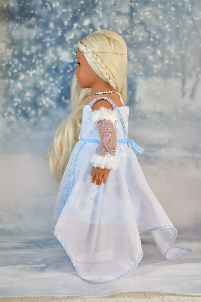 May include: A doll wearing a light blue dress with a white lace overlay and a white fur trim on the sleeve. The doll has long blonde hair and is wearing a silver crown.