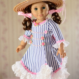 May include: A doll wearing a blue and white striped dress with a pink and white lace trim. The doll is wearing a straw hat with a pink ribbon and a pearl necklace. The doll has brown hair and blue eyes.