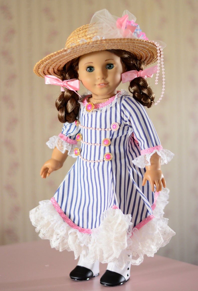 May include: A doll wearing a blue and white striped dress with a pink and white lace trim. The doll is wearing a straw hat with a pink ribbon and a pearl necklace. The doll has brown hair and blue eyes.