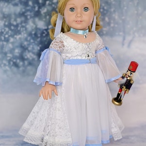 May include: A porcelain doll wearing a white dress with blue accents and a blue ribbon around her neck. She is holding a toy nutcracker in her right hand. The doll is standing on a snowy background.