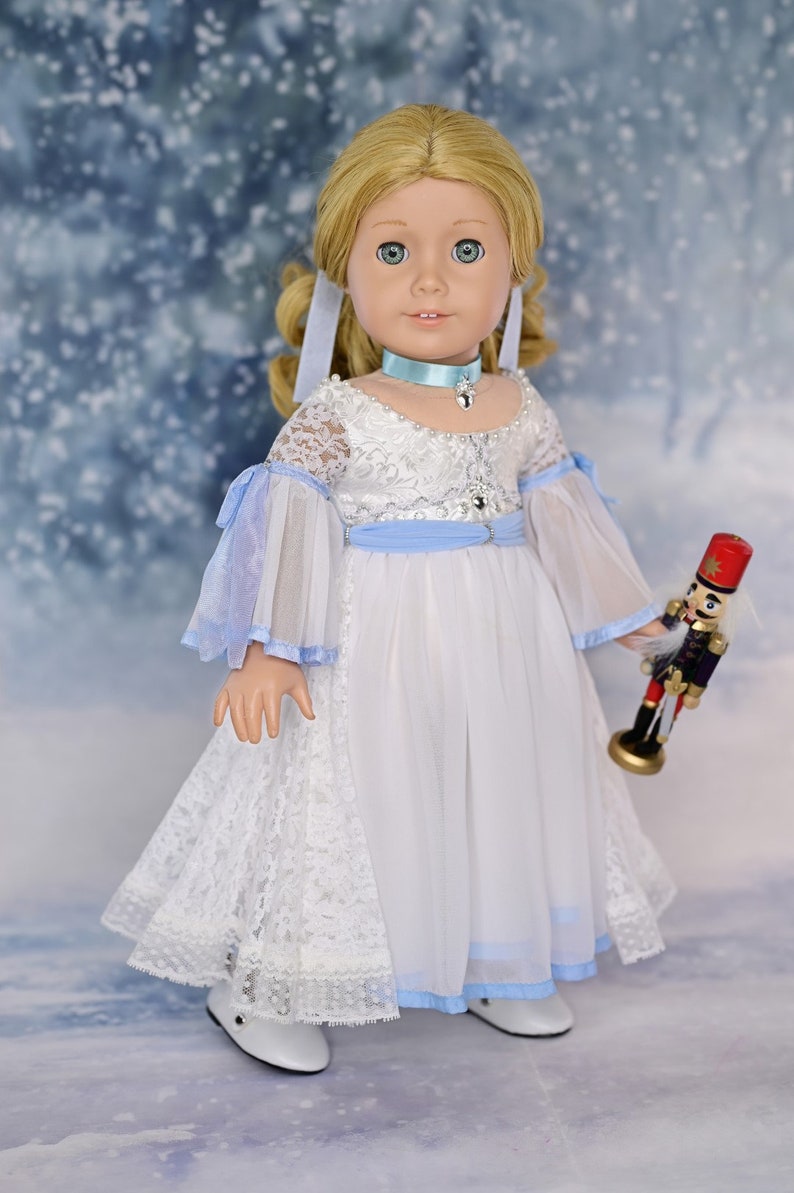 May include: A porcelain doll wearing a white dress with blue accents and a blue ribbon around her neck. She is holding a toy nutcracker in her right hand. The doll is standing on a snowy background.