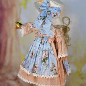 May include: A doll in a light blue floral dress, peach-coloured bodice, and white lace trim. It wears a white hat with a blue floral bow. The doll holds a toy bee. The outfit is reminiscent of a vintage style.