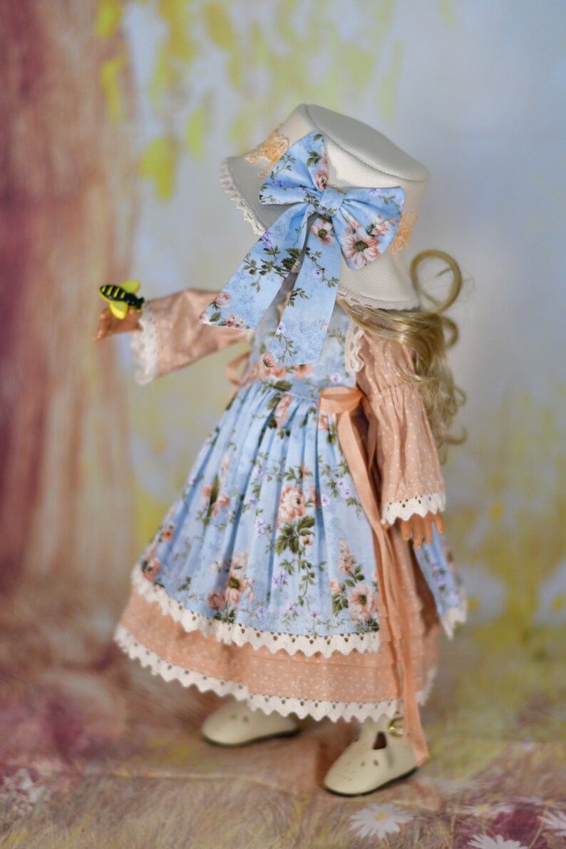 May include: A doll in a light blue floral dress, peach-coloured bodice, and white lace trim. It wears a white hat with a blue floral bow. The doll holds a toy bee. The outfit is reminiscent of a vintage style.