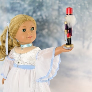 May include: A blonde doll wearing a white dress with blue accents holds a wooden nutcracker toy in her hand. The doll has green eyes and is wearing a blue ribbon around her neck. The background is a blurred image of a snowy forest.