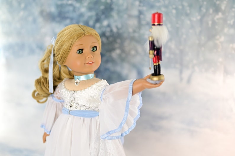 May include: A blonde doll wearing a white dress with blue accents holds a wooden nutcracker toy in her hand. The doll has green eyes and is wearing a blue ribbon around her neck. The background is a blurred image of a snowy forest.