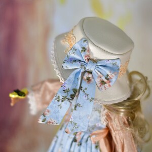 May include: A cream-coloured hat with a light blue floral bow and matching ribbon. The hat has a lace trim and is part of a vintage-inspired outfit. The bow features pink and purple flowers.