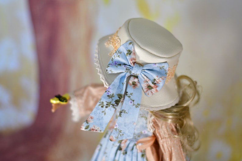May include: A cream-coloured hat with a light blue floral bow and matching ribbon. The hat has a lace trim and is part of a vintage-inspired outfit. The bow features pink and purple flowers.