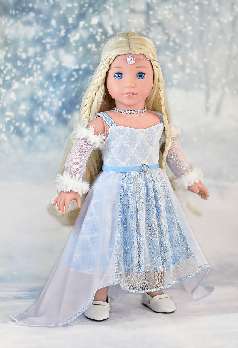 May include: A doll wearing a light blue and white dress with a white fur trim on the sleeves. The doll has long blonde hair and blue eyes. The doll is wearing a silver tiara and a silver necklace. The doll is standing on a white background with a blue and white snowflake pattern.