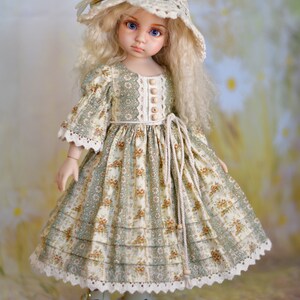 May include: A doll in a cream crocheted hat with a flower and a floral print dress. The dress features a green and cream floral pattern, lace trim, and wooden buttons. The doll is wearing cream shoes.
