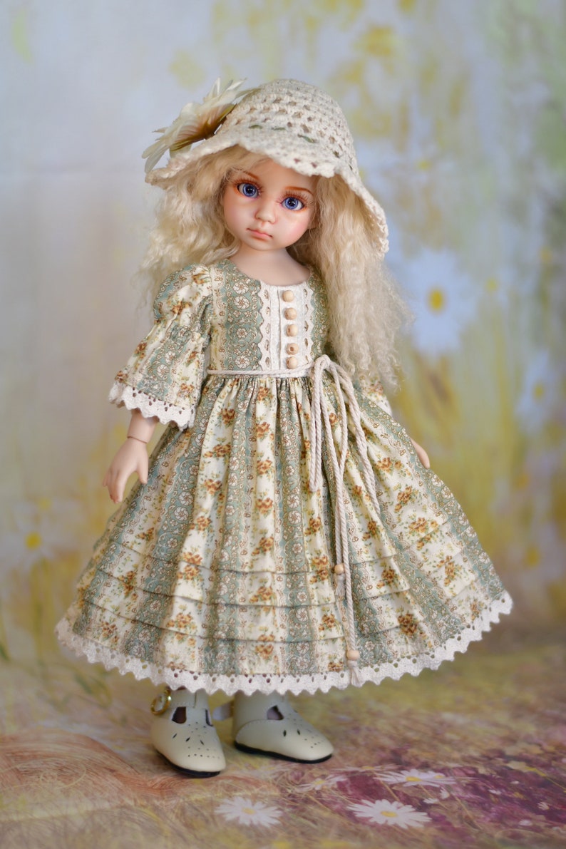 May include: A doll in a cream crocheted hat with a flower and a floral print dress. The dress features a green and cream floral pattern, lace trim, and wooden buttons. The doll is wearing cream shoes.