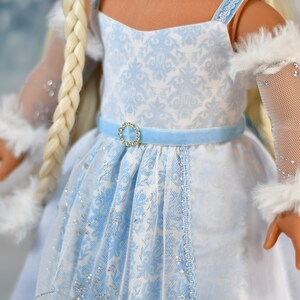 May include: A doll wearing a white and blue dress with a blue velvet sash and a sparkly blue and white lace overlay. The doll has long blonde hair and is wearing white fur-trimmed sleeves with sparkly mesh.