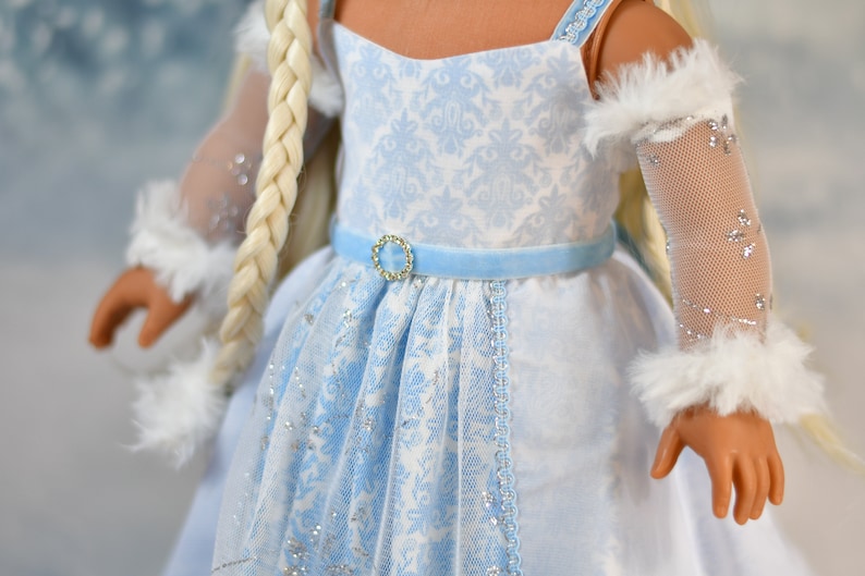 May include: A doll wearing a white and blue dress with a blue velvet sash and a sparkly blue and white lace overlay. The doll has long blonde hair and is wearing white fur-trimmed sleeves with sparkly mesh.