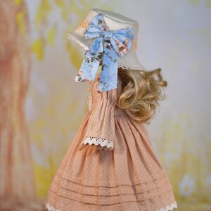 May include: A doll in a peach dress with white polka dots, lace trim, and a matching hat. The hat features a blue floral bow. The doll is wearing white shoes with a buckle.