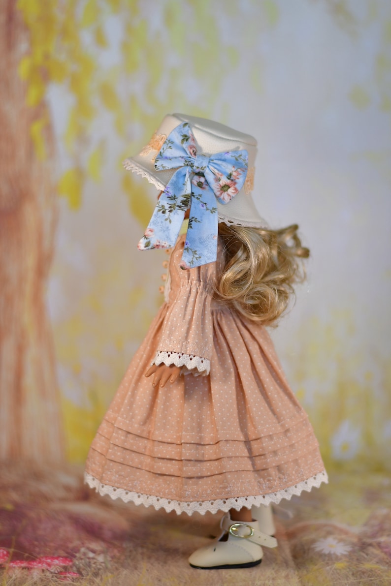 May include: A doll in a peach dress with white polka dots, lace trim, and a matching hat. The hat features a blue floral bow. The doll is wearing white shoes with a buckle.
