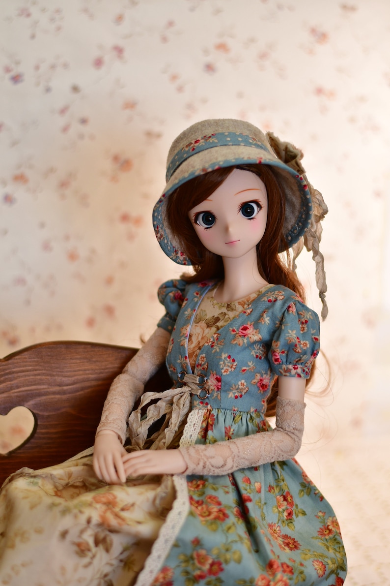 May include: A doll wearing a blue floral dress and a matching hat with a blue ribbon. The doll has long brown hair and is seated on a wooden bench. The dress features a floral pattern with red and pink roses.