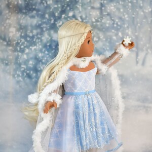 May include: A doll wearing a light blue and white dress with a white fur trim. The doll has long blonde hair and is holding a small snowflake. The doll is standing on a snowy background.