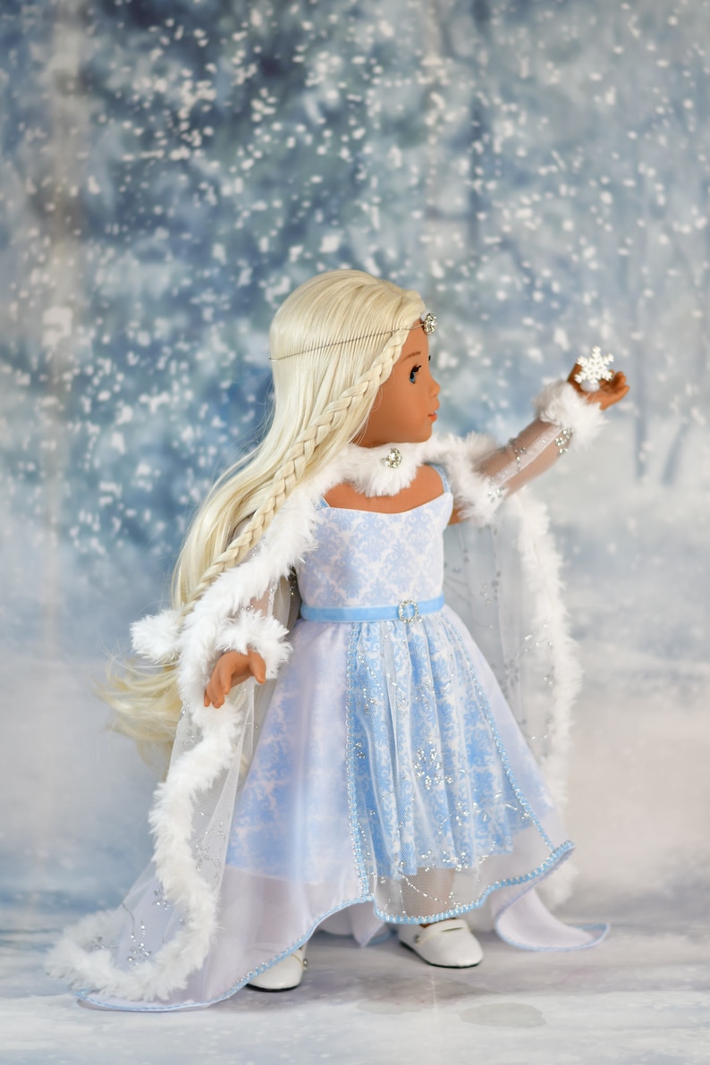 May include: A doll wearing a light blue and white dress with a white fur trim. The doll has long blonde hair and is holding a small snowflake. The doll is standing on a snowy background.