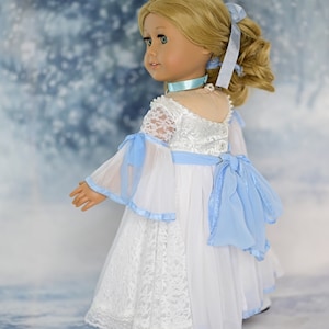 May include: A doll wearing a white lace dress with a blue sash and a blue ribbon in her hair. The doll is standing on a snowy background.