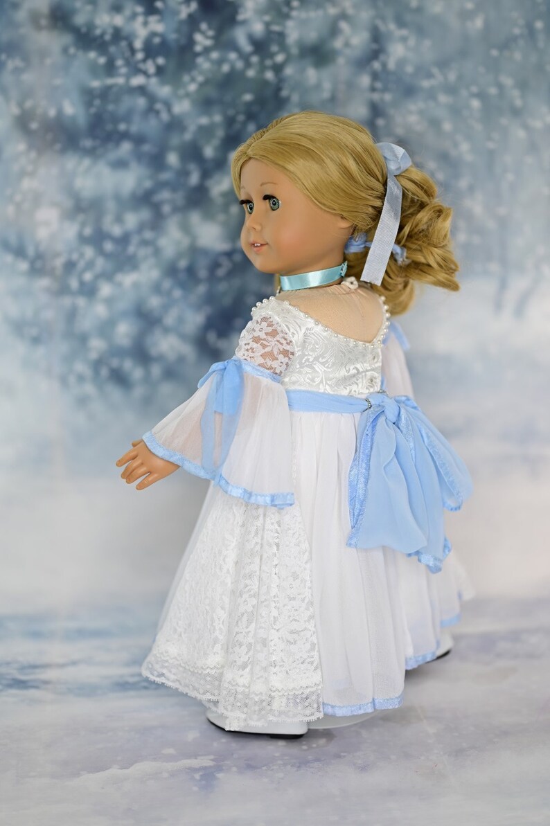 May include: A doll wearing a white lace dress with a blue sash and a blue ribbon in her hair. The doll is standing on a snowy background.