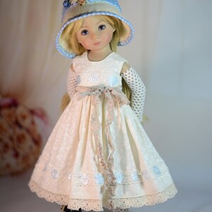 K&ouml;nnte beinhalten: Puppe in einem cremefarbenen Kleid mit floralem Muster, passendem Hut mit blauen Details und schwarzen Schuhen. Das Kleid hat eine Spitzenborte und ein Band um die Taille. Die Puppe hat blondes Haar und blaue Augen.