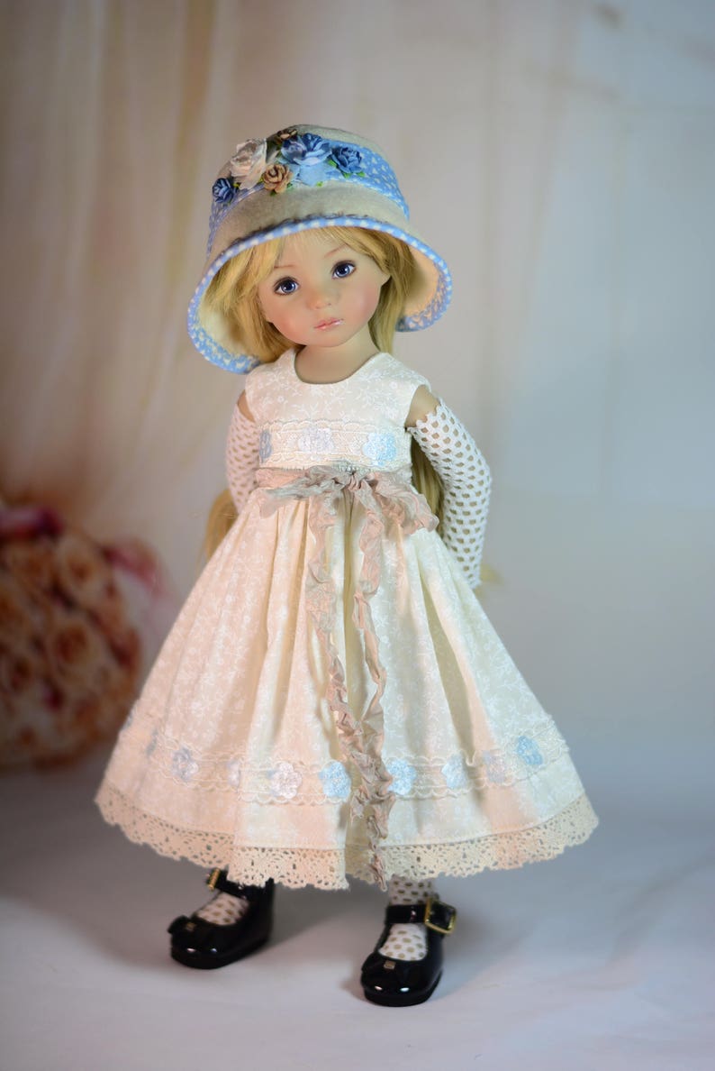 K&ouml;nnte beinhalten: Puppe in einem cremefarbenen Kleid mit floralem Muster, passendem Hut mit blauen Details und schwarzen Schuhen. Das Kleid hat eine Spitzenborte und ein Band um die Taille. Die Puppe hat blondes Haar und blaue Augen.