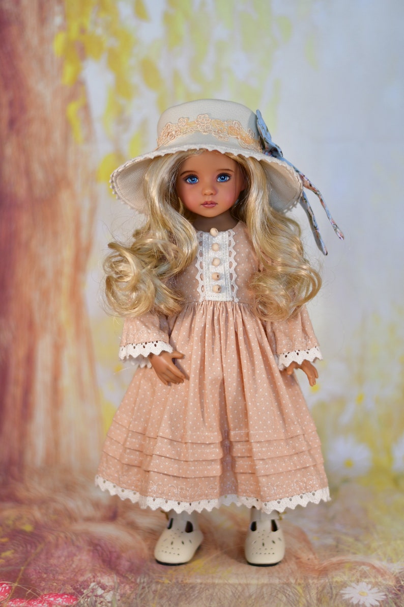 May include: A doll in a peach dress with white polka dots and lace detailing. The doll has long, wavy blonde hair, blue eyes, and a matching hat with a floral bow. The doll is wearing white shoes.