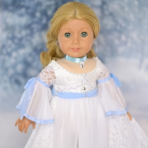 May include: A doll wearing a white dress with blue lace trim and a blue ribbon around the waist. The doll has blonde hair and green eyes. The background is a blurry image of a snowy forest.