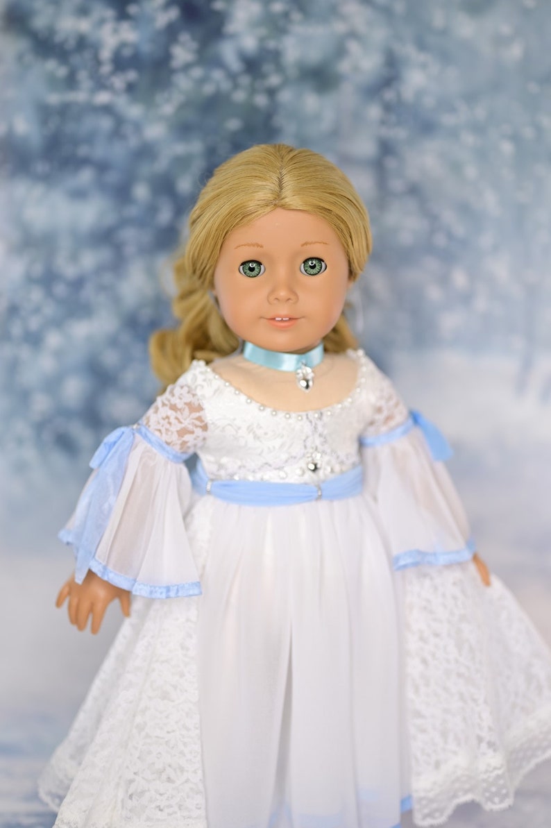 May include: A doll wearing a white dress with blue lace trim and a blue ribbon around the waist. The doll has blonde hair and green eyes. The background is a blurry image of a snowy forest.