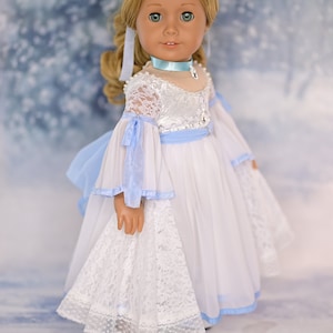 May include: A doll wearing a white dress with blue accents and lace trim. The doll has blonde hair and blue eyes. The doll is wearing white shoes.
