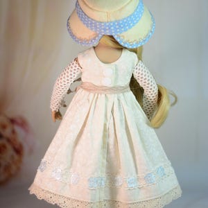K&ouml;nnte beinhalten: Puppe in einem cremefarbenen Kleid mit floralem Muster, Spitzensaum und hellblauen Akzenten. Die Puppe tr&auml;gt einen passenden Hut mit blauem Tupfenband und schwarzen Schuhen. Das Kleid hat einen beigen G&uuml;rtel und wei&szlig;e Kn&ouml;pfe.