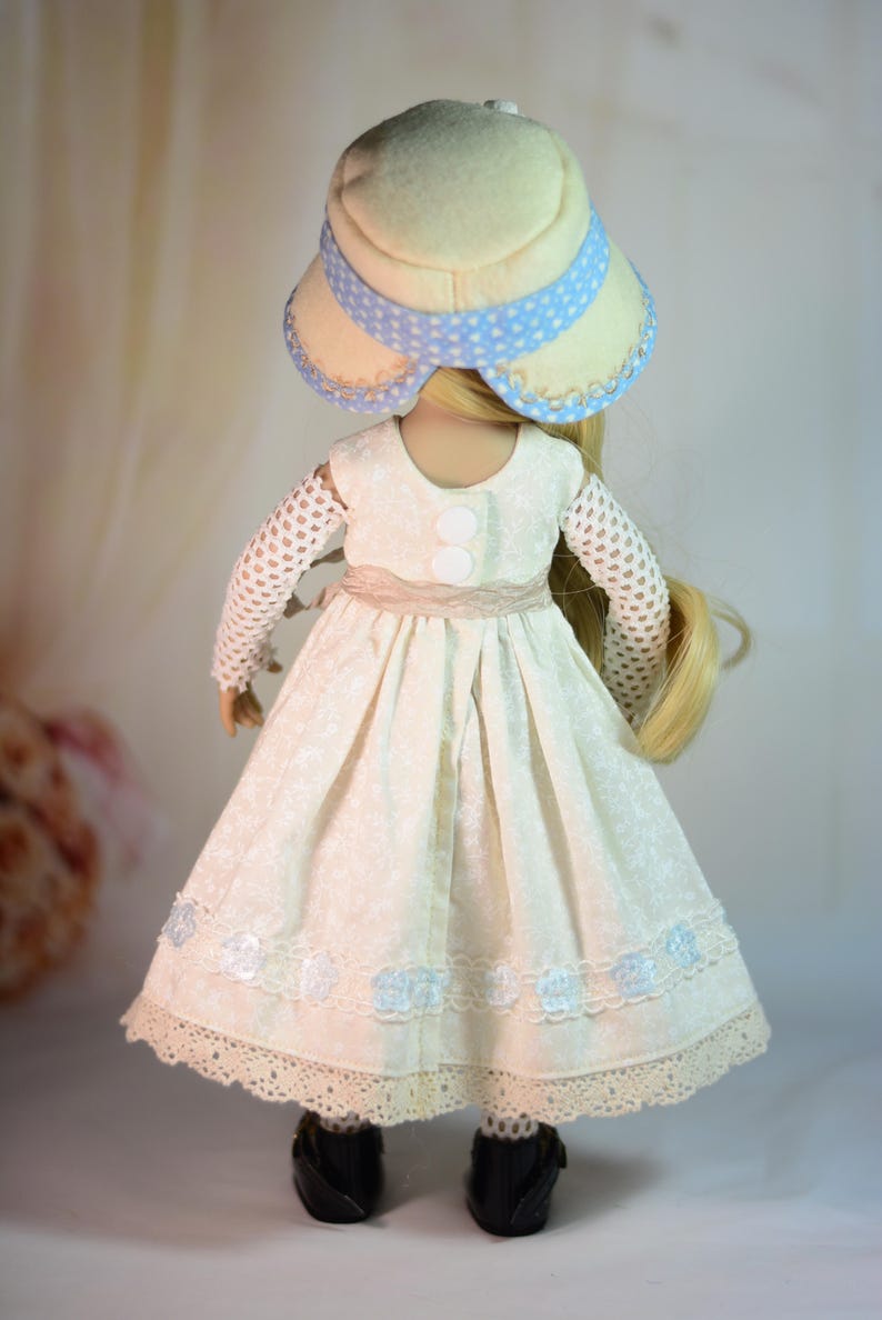 K&ouml;nnte beinhalten: Puppe in einem cremefarbenen Kleid mit floralem Muster, Spitzensaum und hellblauen Akzenten. Die Puppe tr&auml;gt einen passenden Hut mit blauem Tupfenband und schwarzen Schuhen. Das Kleid hat einen beigen G&uuml;rtel und wei&szlig;e Kn&ouml;pfe.