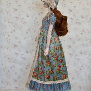 May include: A doll wearing a vintage-style dress with a floral pattern in red and orange on a blue background. The doll also wears a matching hat with a floral accent and brown shoes. The dress has lace trim and long, sheer sleeves.