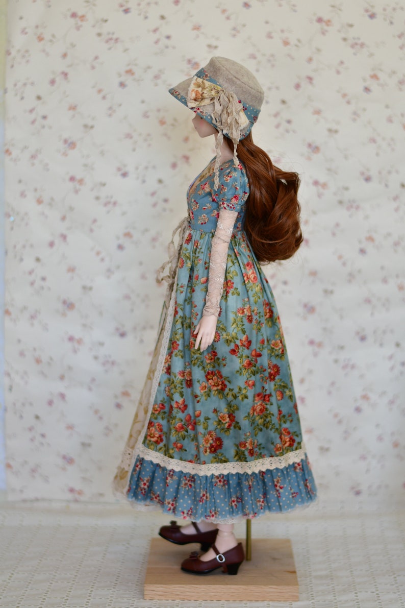 May include: A doll wearing a vintage-style dress with a floral pattern in red and orange on a blue background. The doll also wears a matching hat with a floral accent and brown shoes. The dress has lace trim and long, sheer sleeves.
