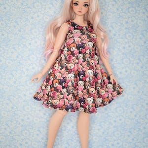 May include: A doll wearing a pink and black dress with a teddy bear and rose print. The doll has long pink hair and is wearing white shoes.