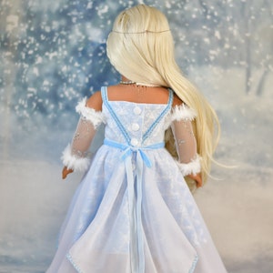 May include: A doll wearing a light blue dress with a white snowflake pattern. The dress has a white fur trim on the sleeves and a light blue ribbon tied at the waist. The doll has long blonde hair and is standing in front of a snowy background.