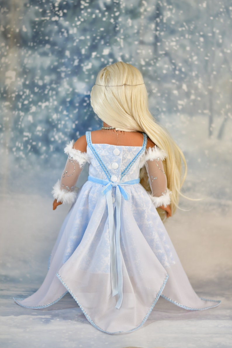 May include: A doll wearing a light blue dress with a white snowflake pattern. The dress has a white fur trim on the sleeves and a light blue ribbon tied at the waist. The doll has long blonde hair and is standing in front of a snowy background.