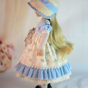 K&ouml;nnte beinhalten: Eine Puppe in einem Kleid und Hut im Vintage-Stil. Das Kleid ist cremefarben mit blauen Blumenmustern und einem R&uuml;schensaum. Der Hut ist cremefarben mit einem blauen Tupfenband. Die Puppe hat lange blonde Haare und schwarze Schuhe.