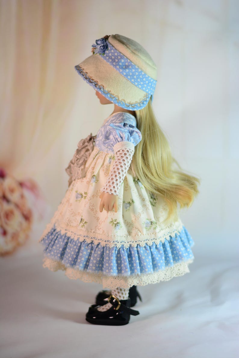 K&ouml;nnte beinhalten: Eine Puppe in einem Kleid und Hut im Vintage-Stil. Das Kleid ist cremefarben mit blauen Blumenmustern und einem R&uuml;schensaum. Der Hut ist cremefarben mit einem blauen Tupfenband. Die Puppe hat lange blonde Haare und schwarze Schuhe.