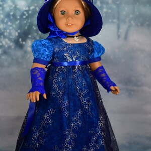 May include: A blue and silver doll's dress with a matching blue hat and gloves. The dress has a floral pattern and a satin sash. The doll is wearing a silver necklace.
